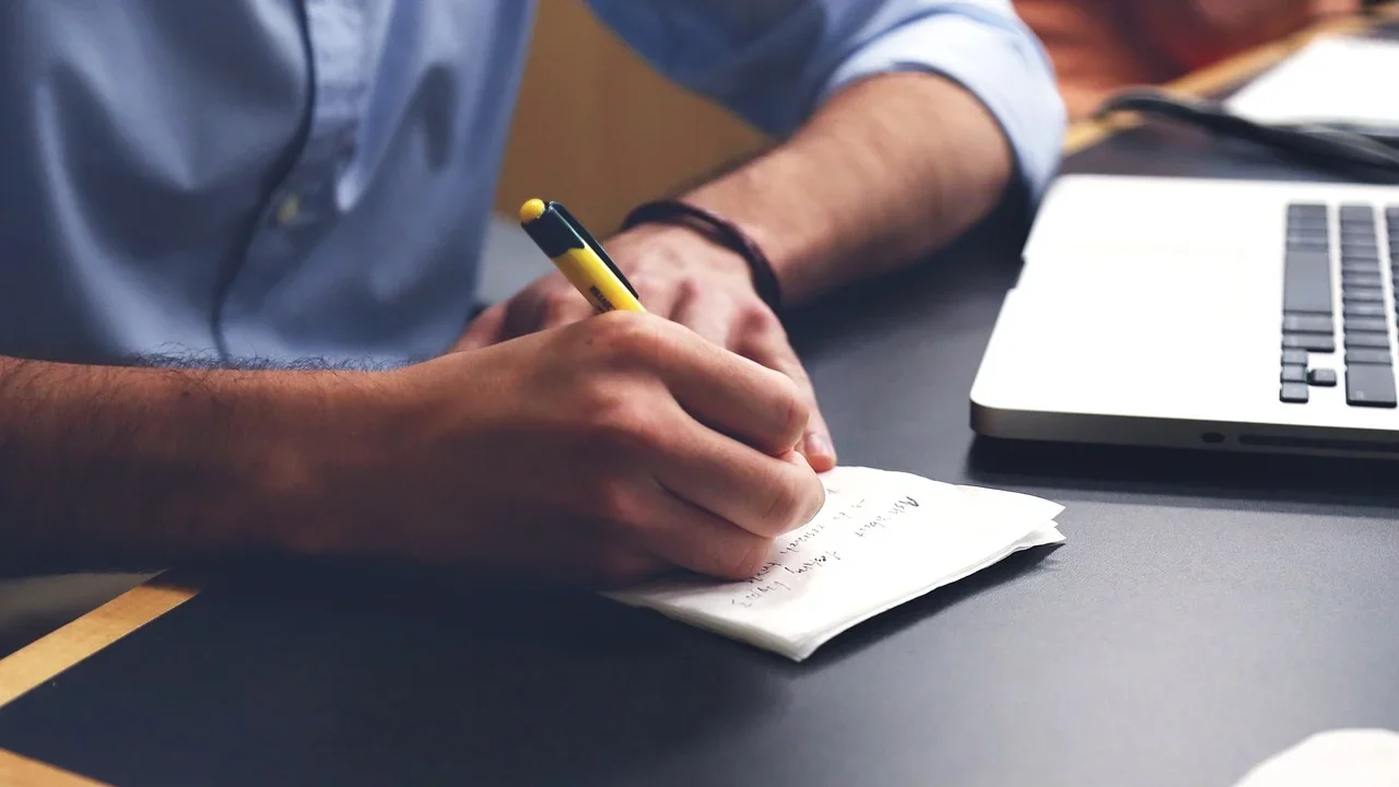 Man jotting down notes on a notepad with a computer next to him.