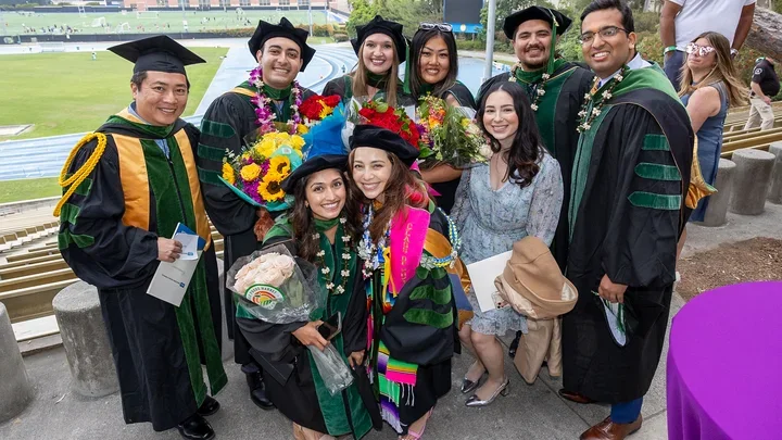 A group of graduates in caps and gowns smiling together at DGSOM Commencement ceremony, celebrating their achievements.