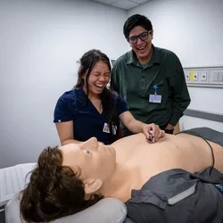 A cheerful scene featuring two individuals smiling at a mannequin, highlighting their enjoyment and engagement with the mannequin. simulating a medical exam. 